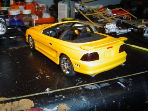 1979 Ford Bronco - A yellow convertible model car sits on a cluttered workbench, surrounded by hobby supplies and other unfinished models.