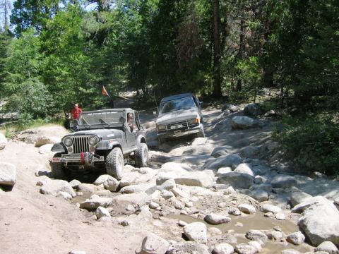 1985 Ford Bronco - Two off-road vehicles, a Jeep and a Ford Bronco, tackle a challenging rocky path in a wooded area.