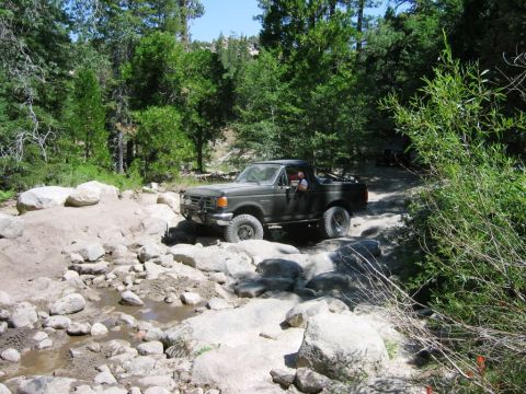 1985 Ford Bronco - A dark-colored Ford Bronco drives through a rocky stream bed on a wooded trail.