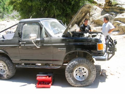 1985 Ford Bronco - Two men work on a black 1985 Ford Bronco with its hood open in an outdoor, rocky area.