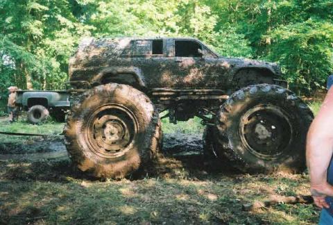 1987 Suzuki Samurai - This heavily modified SUV, covered in mud, sits low with massive tires, while a pickup truck is partially visible in the background.
