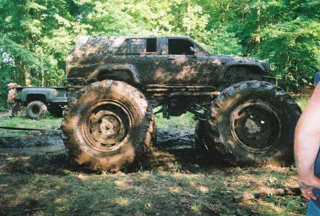 Mud-covered SUV with oversized tires, side view. Another truck is visible behind it