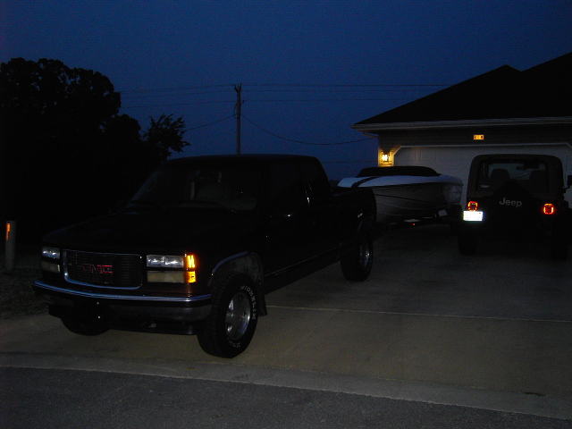 Black GMC pickup truck with a boat trailer, parked in a driveway at dusk. A Jeep is visible to the right