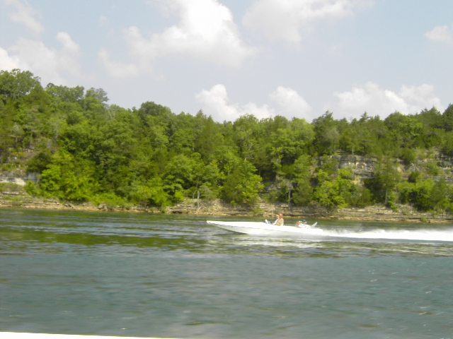 White speedboat with two people speeds across a lake with tree-covered cliffs in the background