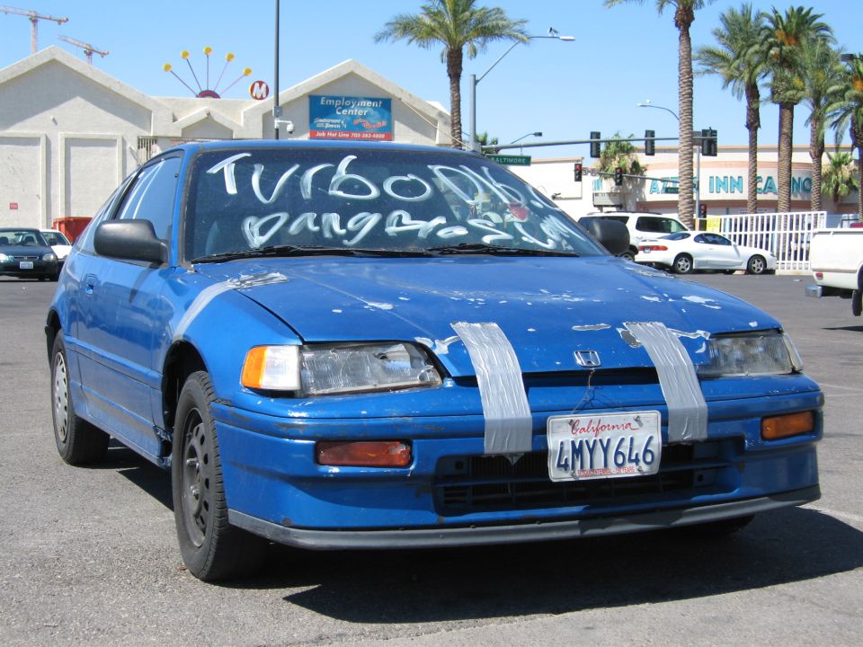 Blue Honda CRX with duct-taped hood and windshield writing in parking lot