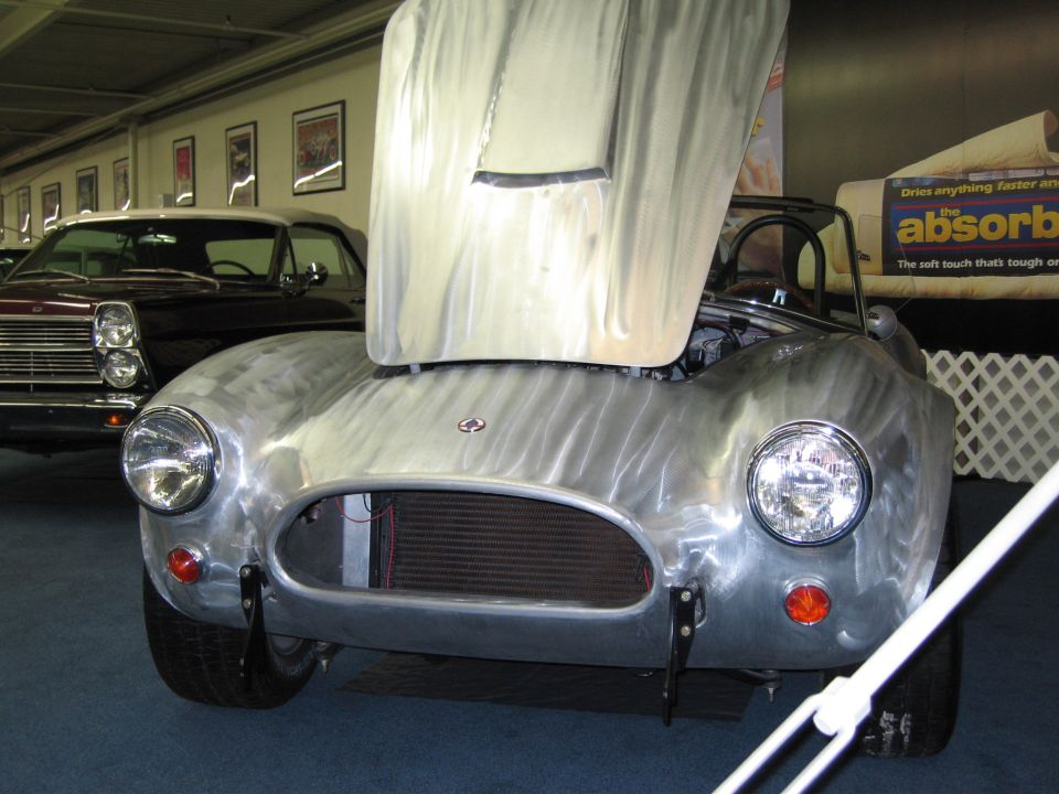 Aluminum Shelby Cobra with hood open, front view. Another car is visible behind it