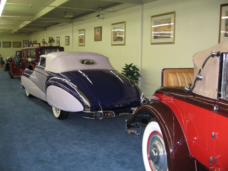 Two classic convertible cars parked indoors, one dark blue and light purple, the other red and dark brown