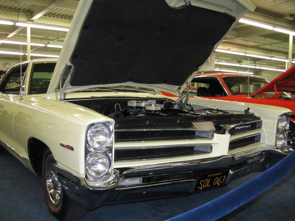 Front view of a cream-colored 1965 Pontiac with hood open, showing engine. Red car visible behind