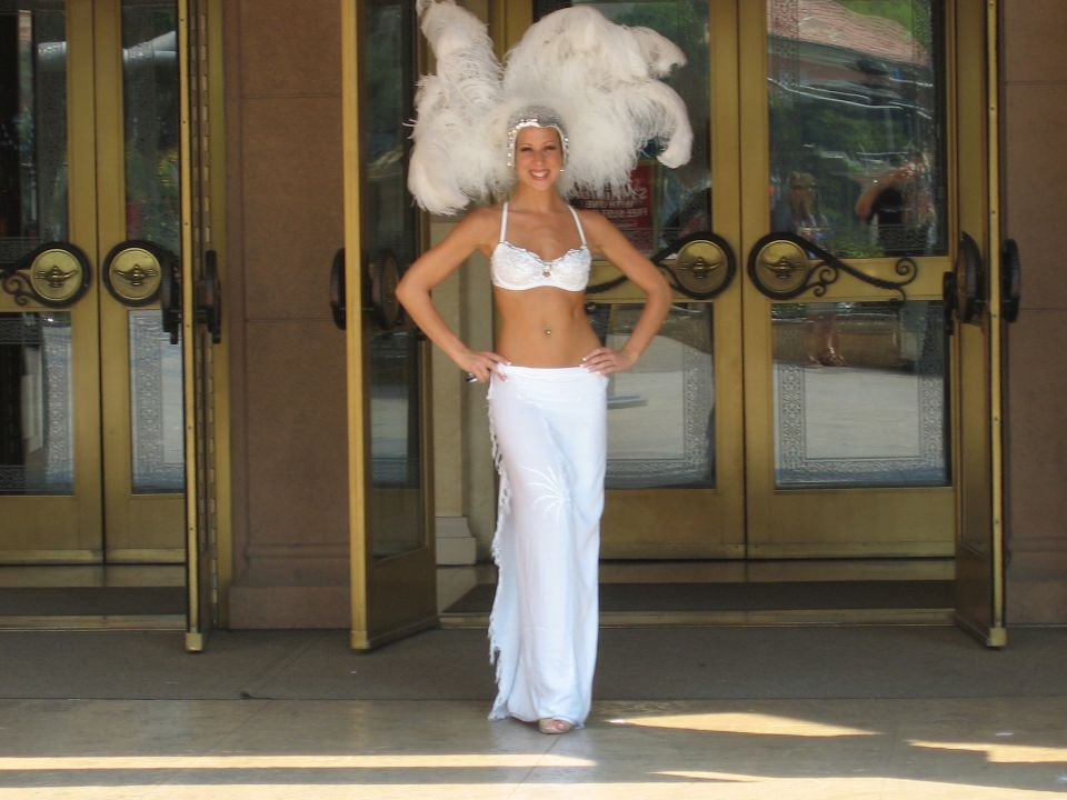 Woman in white costume with feather headdress stands before double doors