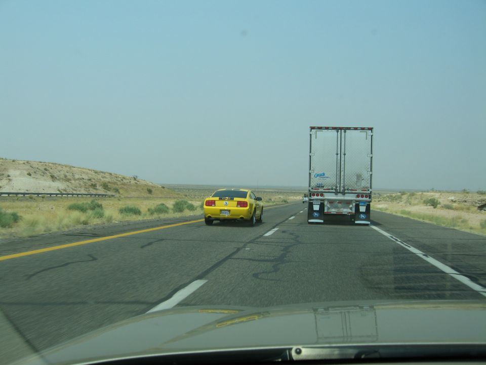 Yellow Ford Mustang driving on highway behind a semi-trailer truck. The Mustang appears to be a 2005 model
