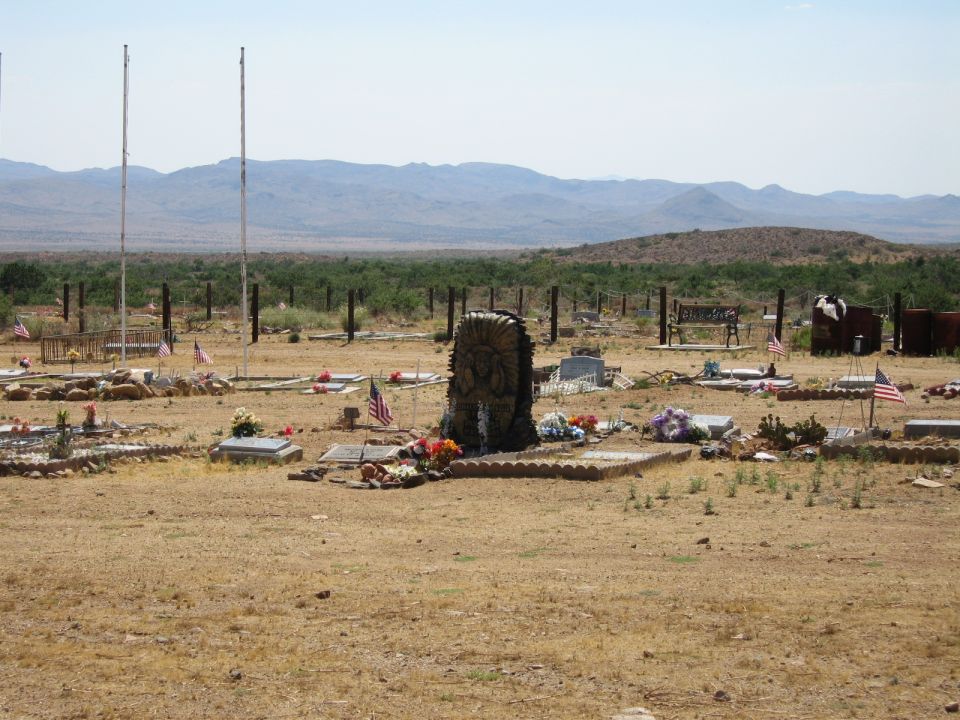 Desert cemetery with graves, flags, and a chief-carved headstone, mountains behind.