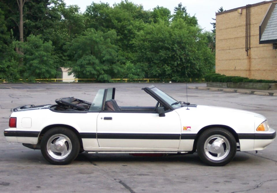 White Ford Mustang convertible, top down, side view in a parking lot with trees and brick building behind