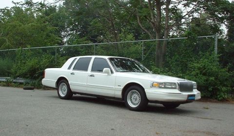 1997 Ford Crown Victoria - This white Lincoln Town Car is parked on a paved surface with a chain-link fence and trees in the background.