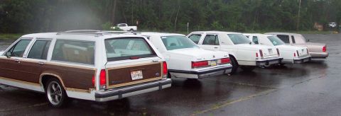 1985 Ford LTD Crown Victoria - A line of cars, including a wood-paneled station wagon, are parked on a wet asphalt surface.