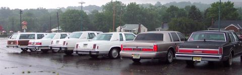1985 Ford LTD Crown Victoria - A row of sedans and station wagons, including a beige Lincoln Town Car, are parked in a lot on a rainy day.