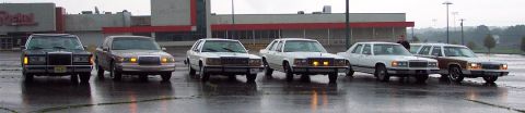 1985 Ford LTD Crown Victoria - Several Ford sedans, including a wood-paneled wagon, are parked in a lot on a rainy day.