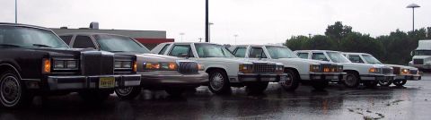 1985 Ford LTD Crown Victoria - Several sedans are parked in a line on a wet lot under an overcast sky.