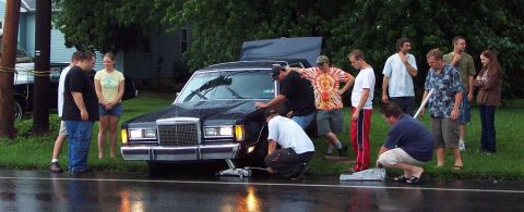 1985 Ford LTD Crown Victoria - A group of people attend to a black sedan with its hood up and a jack positioned under the front wheel on a wet road.