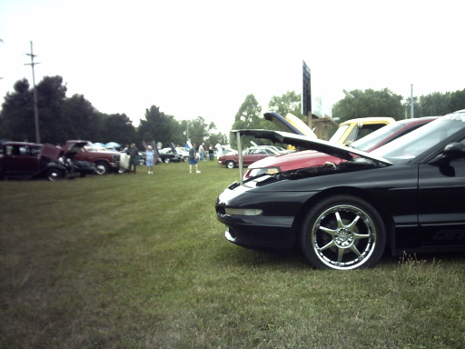 Black 1995 Ford Probe with hood open, parked on grass at a car show. Other cars and people are visible in the background