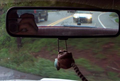 1985 Ford LTD Crown Victoria - A white car is seen in the rearview mirror, driving on a wet road with another vehicle ahead.