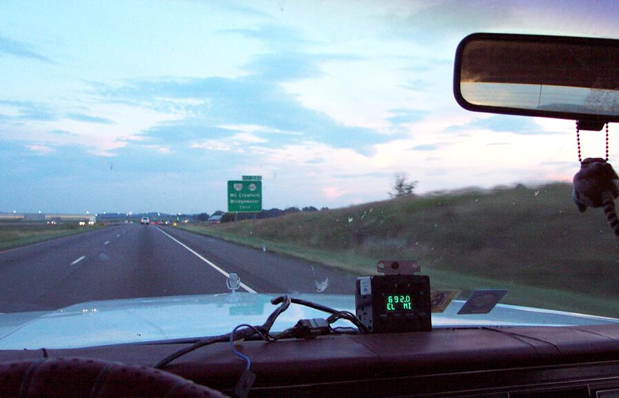 Dashboard view from inside a car on a highway, with a radar detector showing '6920 EL MI'