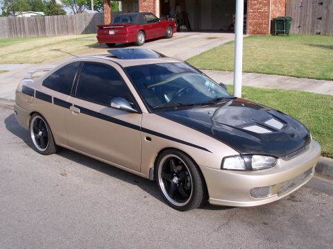 1999 Mitsubishi Mirage - This tan coupe, identified as a 1999 Mitsubishi Mirage, features black racing stripes and an aftermarket hood.
