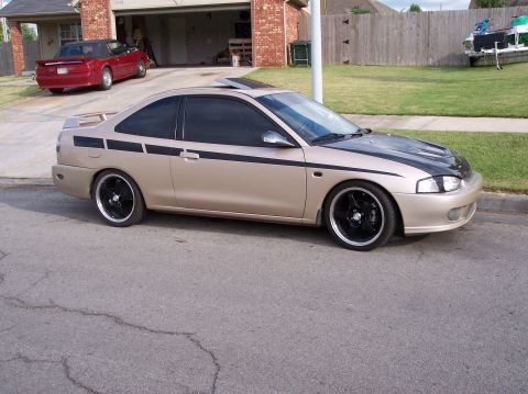 1999 Mitsubishi Mirage - Tan 1999 Mitsubishi Mirage coupe with black racing stripes and aftermarket wheels is parked in a driveway.