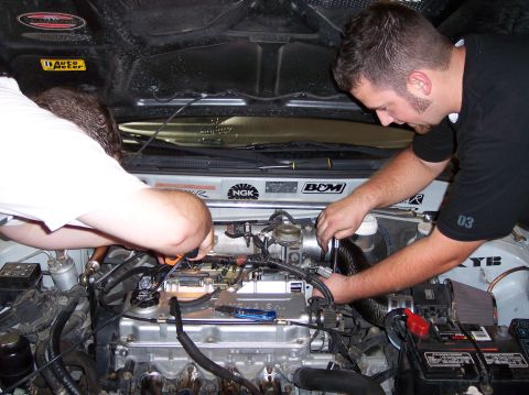 1999 Mitsubishi Mirage - Two mechanics are actively working on the engine of a car, with the hood open and tools visible.