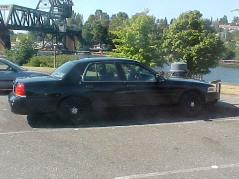 1999 Ford Crown Victoria - Side view of a black 1999 Ford Crown Victoria sedan parked outdoors near a body of water and a bridge.