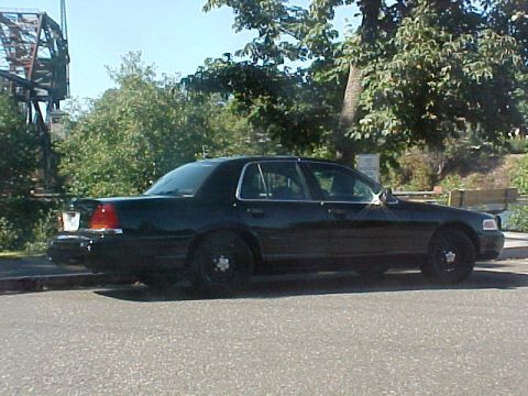 1999 Ford Crown Victoria - A black sedan is parked on the side of the road with trees and a bridge visible.