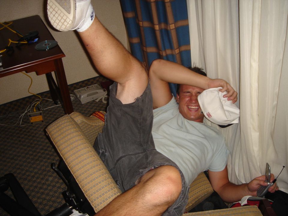 Man laughing in armchair holding a white cap and flip phone indoors