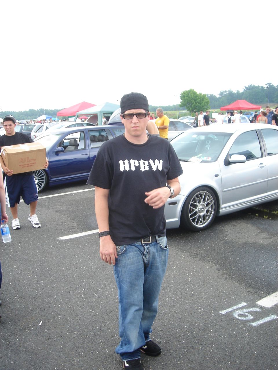 Man in black HPBW shirt standing near silver VW Jetta sedan at a car meet