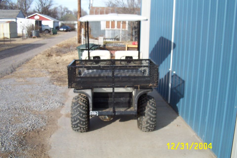 Rear of small utility golf cart with white roof and mesh cargo bed by a blue building