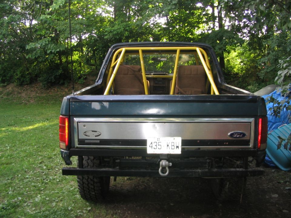 Rear view of a dark green 1986 Ford Bronco with a yellow roll cage and a silver tailgate panel