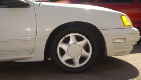 1991 Ford Taurus - Close-up of the front fender and wheel of a white 1991 Ford Taurus SHO, with another vehicle visible behind it.