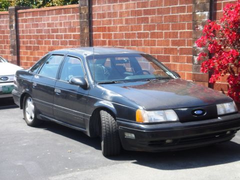 1991 Ford Taurus - A dark 1991 Ford Taurus is parked in front of a brick wall, with another vehicle partially visible to the left.