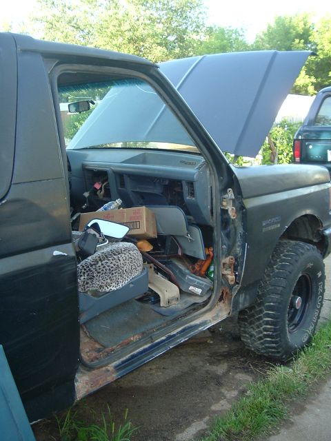 1989 Ford Bronco - The interior of a Ford Bronco is visible with the driver's side door open and the hood raised, revealing a cluttered dashboard and seat.