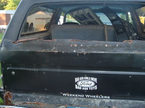1989 Ford Bronco - This vehicle's rear shows significant rust and custom stickers, including one that says 'Bad Ass Girls Drive Ford Bad Ass Toys'.