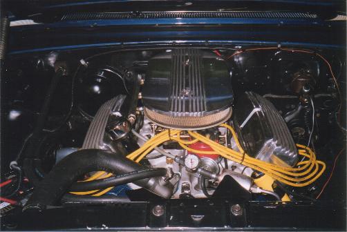 V8 engine bay with finned air cleaner, finned valve covers, and yellow spark plug wires in a dark blue car