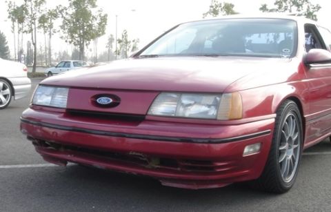 1991 Ford Taurus - Front three-quarter close-up of a red first-generation Ford Taurus SHO with a scraped and cracked lower front bumper cover showing bare dam…