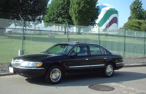 1997 Ford Crown Victoria - A black sedan with chrome accents is parked outdoors near a chain-link fence and a large, colorful cylindrical structure.