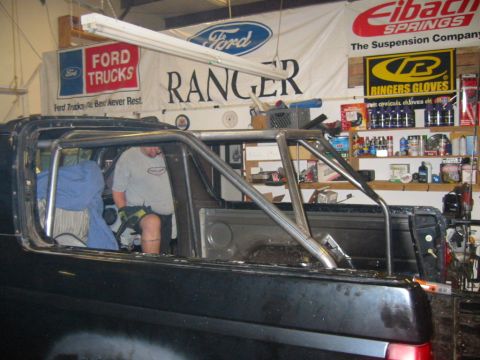 1992 Ford Bronco - Side view of a black truck cab stripped of glass and interior trim with a bare bent-tube roll cage being fabricated inside, a man in a grey…