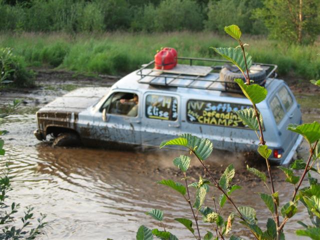 Silver SUV stuck in mud with text on side: 'NV Softba Defendin CHAMPS'