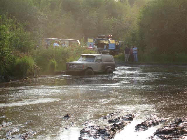 Tan SUV navigating a muddy water crossing with other vehicles and people in the background