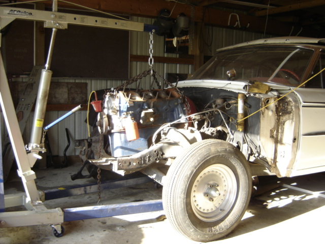 V8 engine on a hoist being pulled from a stripped white full-size Ford sedan in a garage
