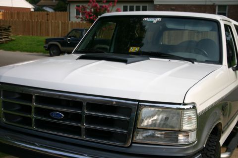 1994 Ford Bronco - Front view of a white Ford Bronco with a hood scoop, showing the grille and headlights. Another truck is visible in the background.