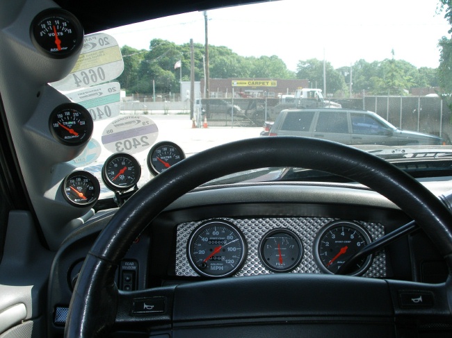 Car interior with black steering wheel and multiple aftermarket Auto Meter gauges on dash and A-pillar.