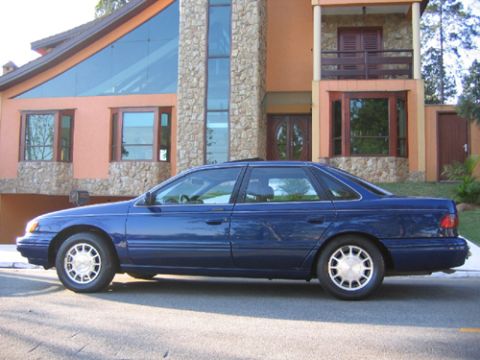 1995 Ford Taurus - A blue 1995 Ford Taurus sedan is parked on a street in front of a large, multi-story house.