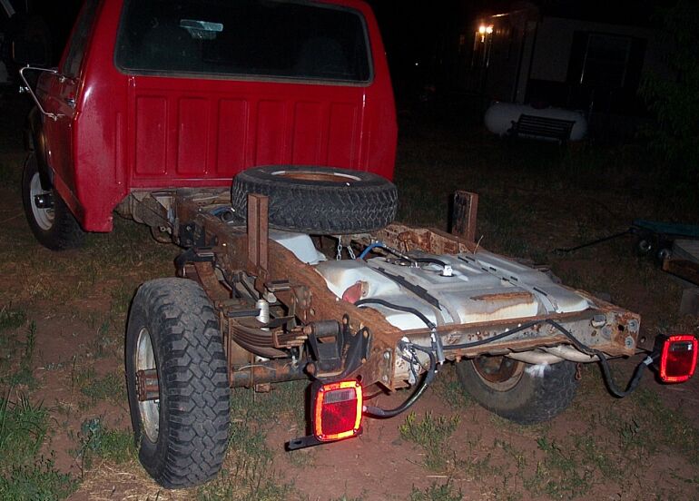Red truck chassis missing body, with spare tire mounted above fuel tank. Rear view shows leaf springs and taillights