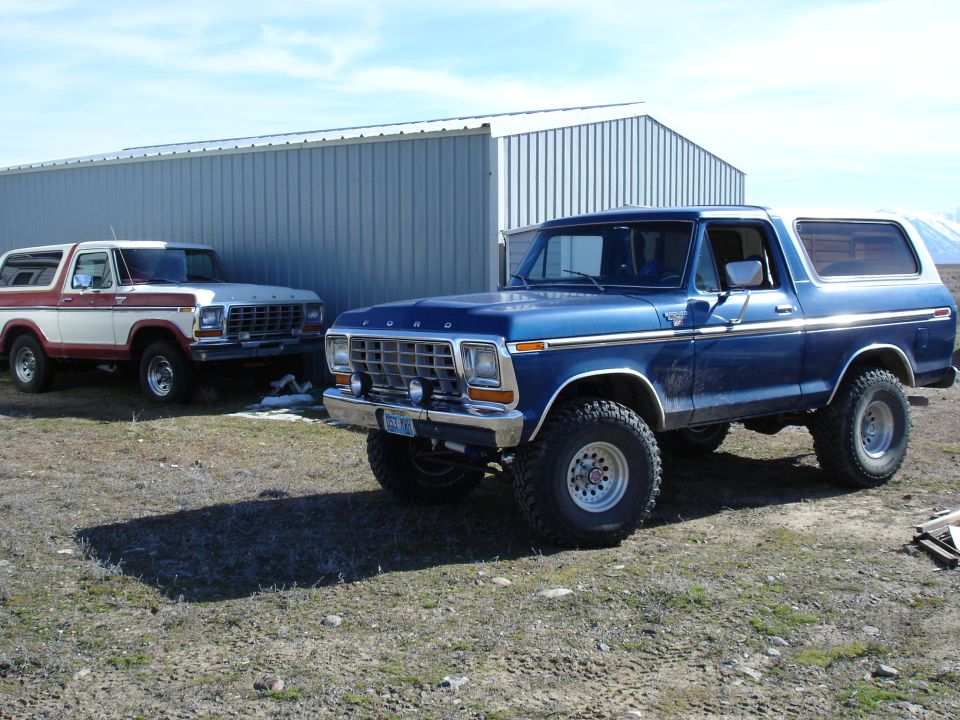 Two Ford Broncos parked outside a metal building. The blue one is in the foreground